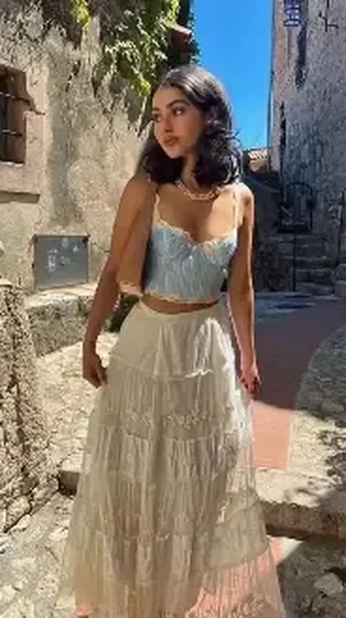 Woman in a pale blue crochet crop top and cream tiered fringed maxi skirt with a shell necklace in a sunlit Mediterranean stone alley