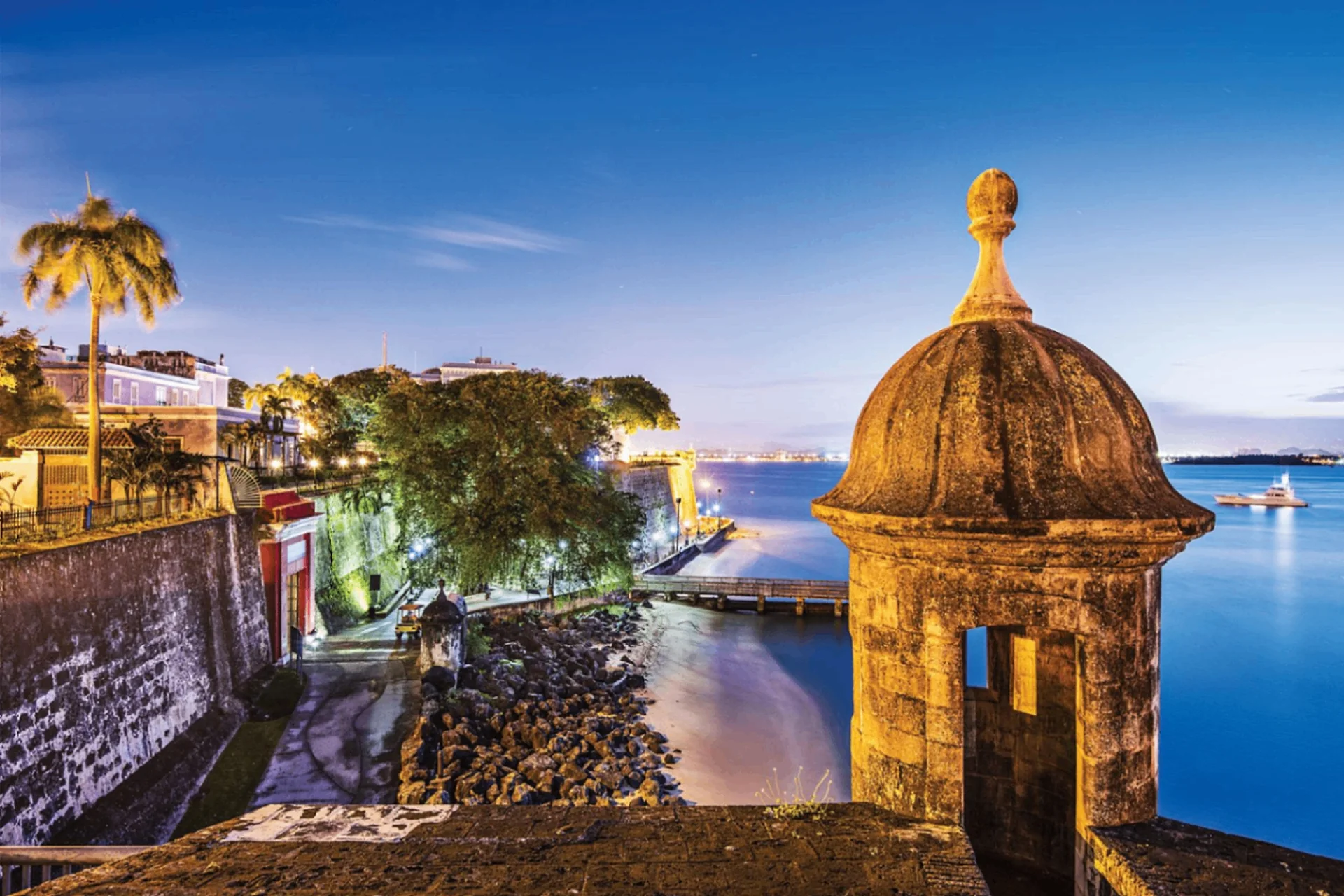 Old San Juan at twilight with a lit stone sentry box in the foreground, the colonial city wall, palm trees, lit buildings, and a calm blue bay