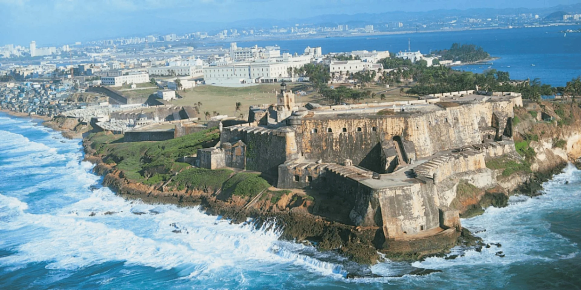 Aerial view of El Morro fort on its headland with green lawns, the walled city and modern skyline behind, and white surf breaking against the rocks below