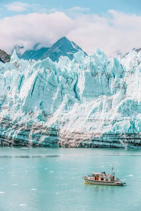 A small tour boat on pale turquoise water drifting before the towering blue-white face of a tidewater glacier beneath low clouds